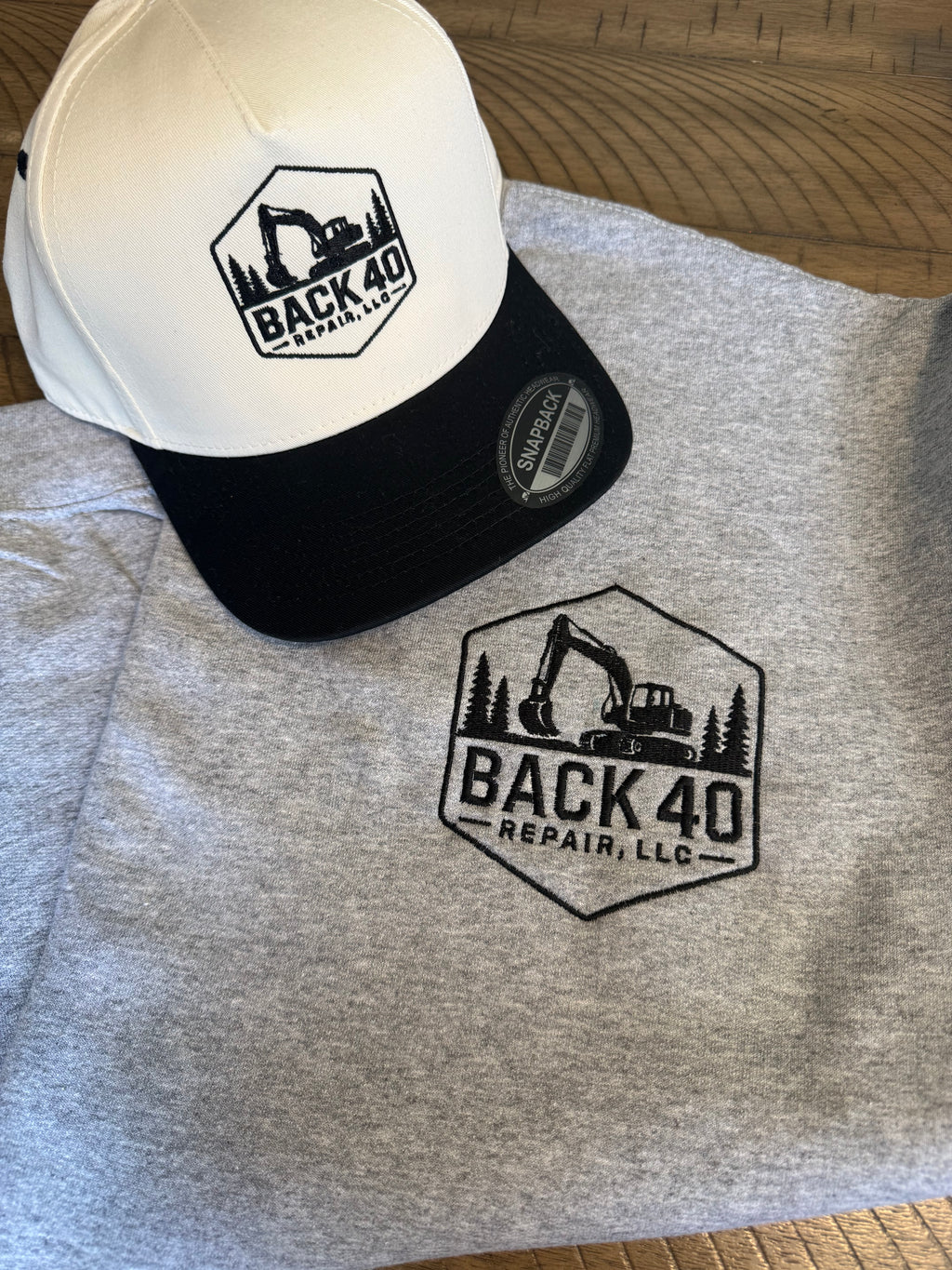 Gray crew neck sweatshirt and white and black cap with 'Back 40 Repair, LLC' logo on a wooden surface.