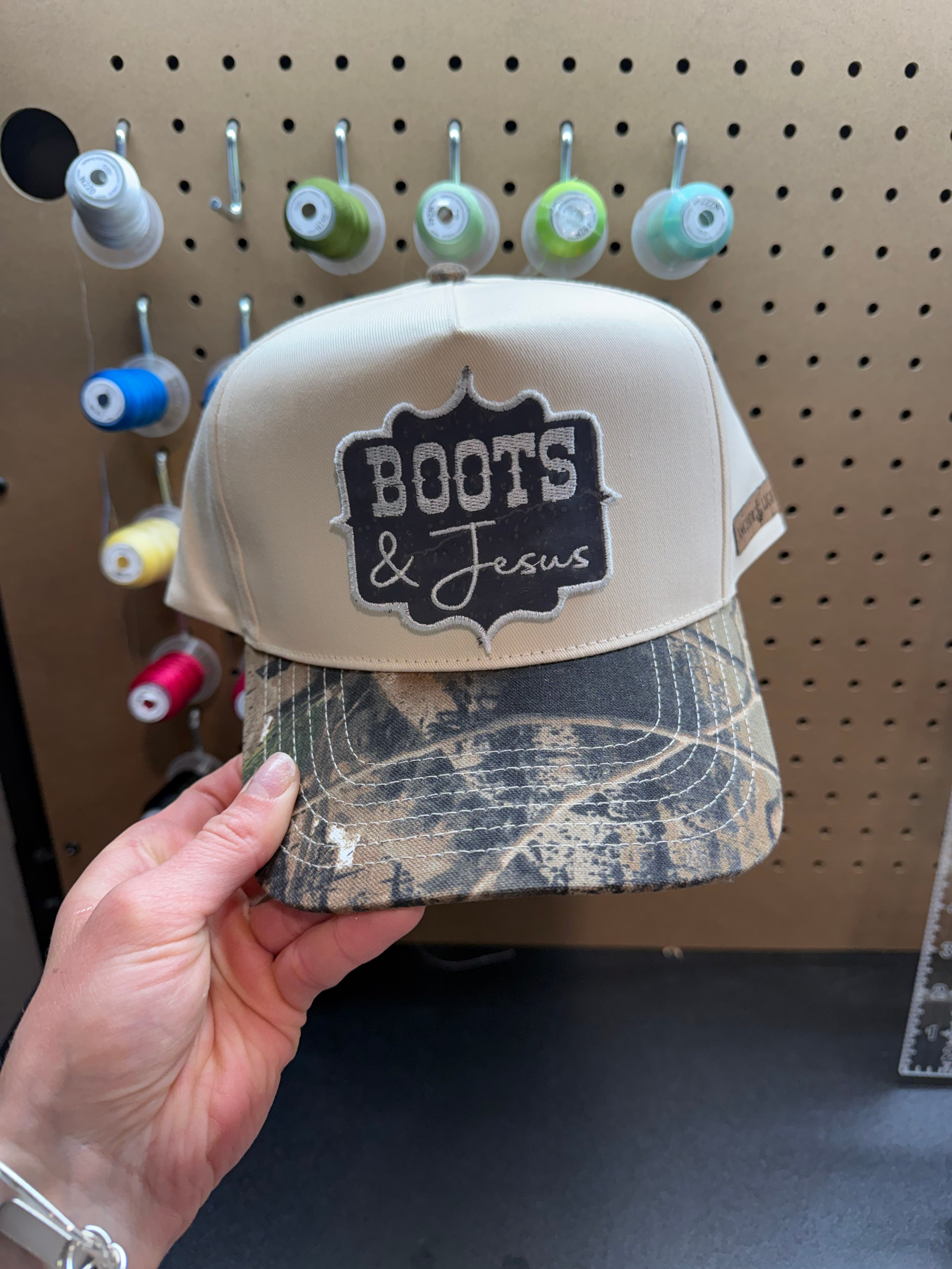 Camouflage-patterned cap with 'Boots & Jesus' logo held by a hand against a pegboard background.