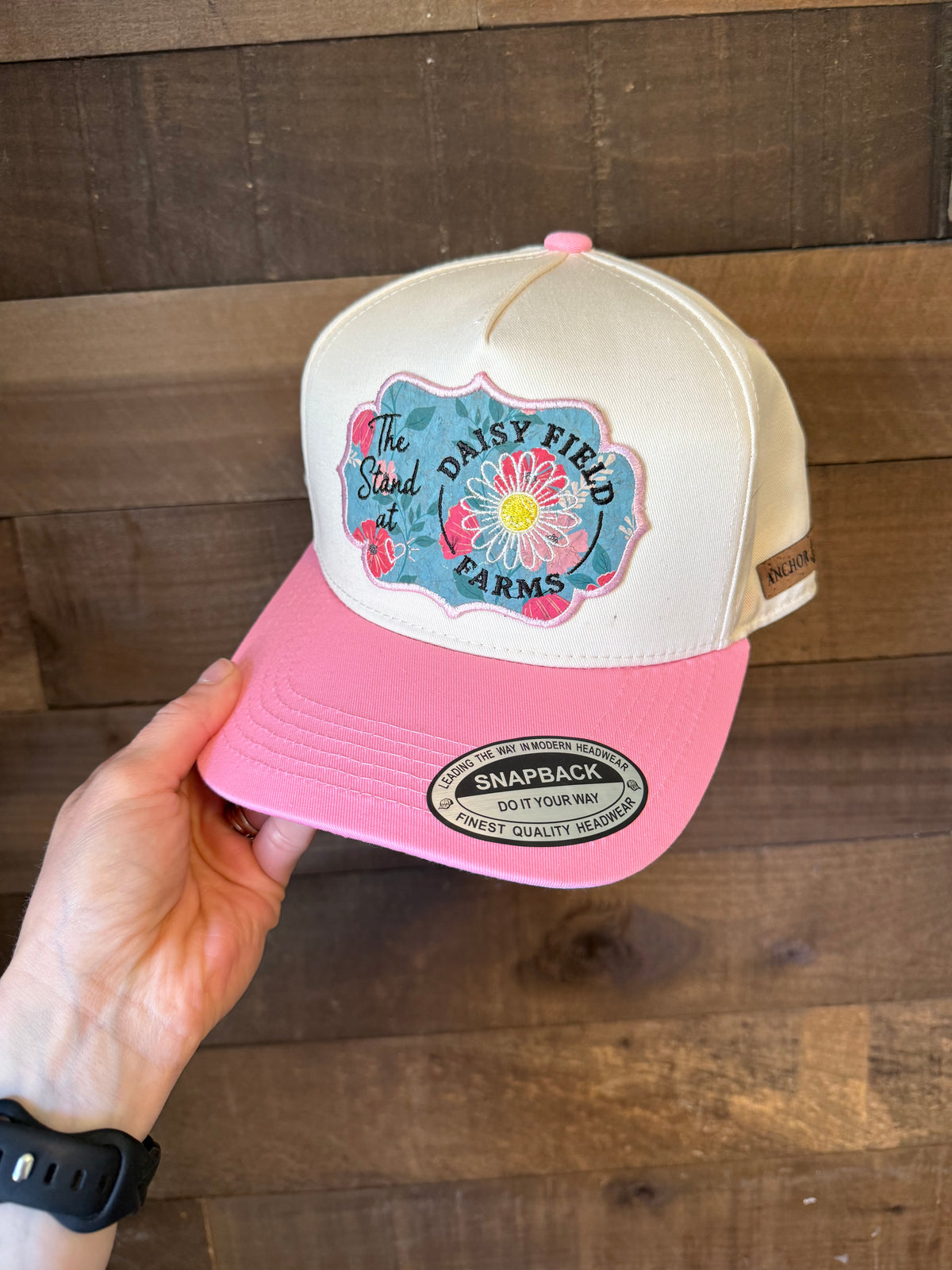 Pink and beige cap with 'Daisy Field Farms' logo held against a wooden background