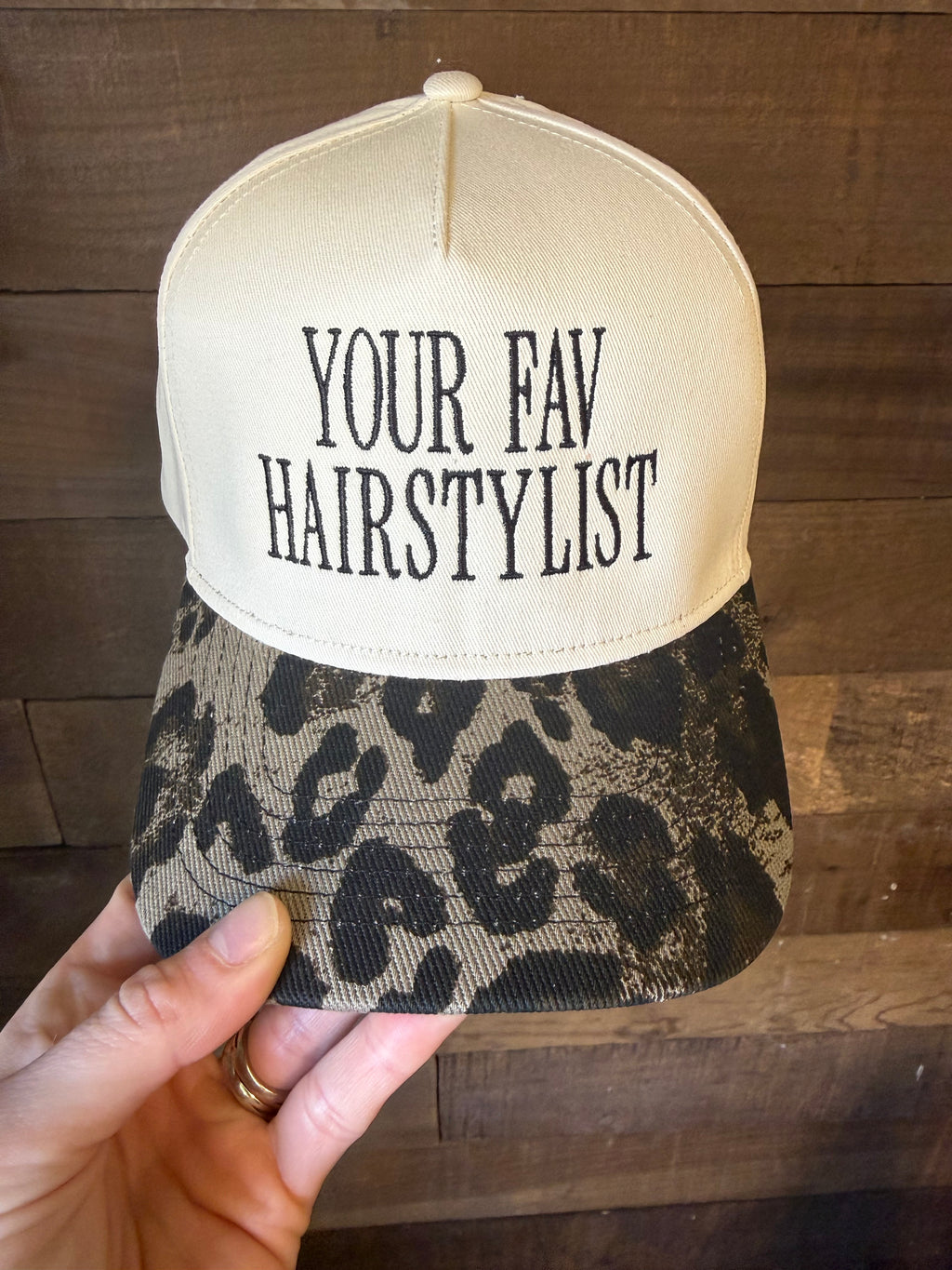 Cap with 'YOUR FAV HAIRSTYLIST' text held by a hand on a wooden surface