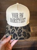 Cap with 'YOUR FAV HAIRSTYLIST' text held by a hand on a wooden surface