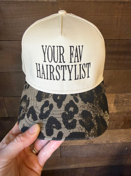 Cap with 'YOUR FAV HAIRSTYLIST' text held by a hand on a wooden surface