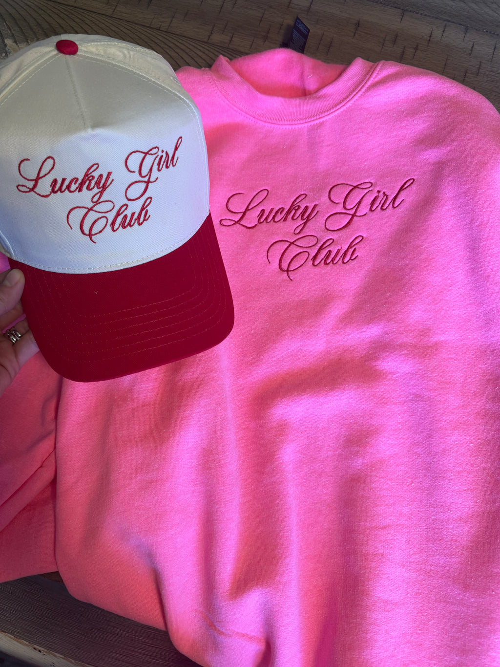 Pink sweatshirt and red and white cap with 'Lucky Girl Club' text on a wooden surface.