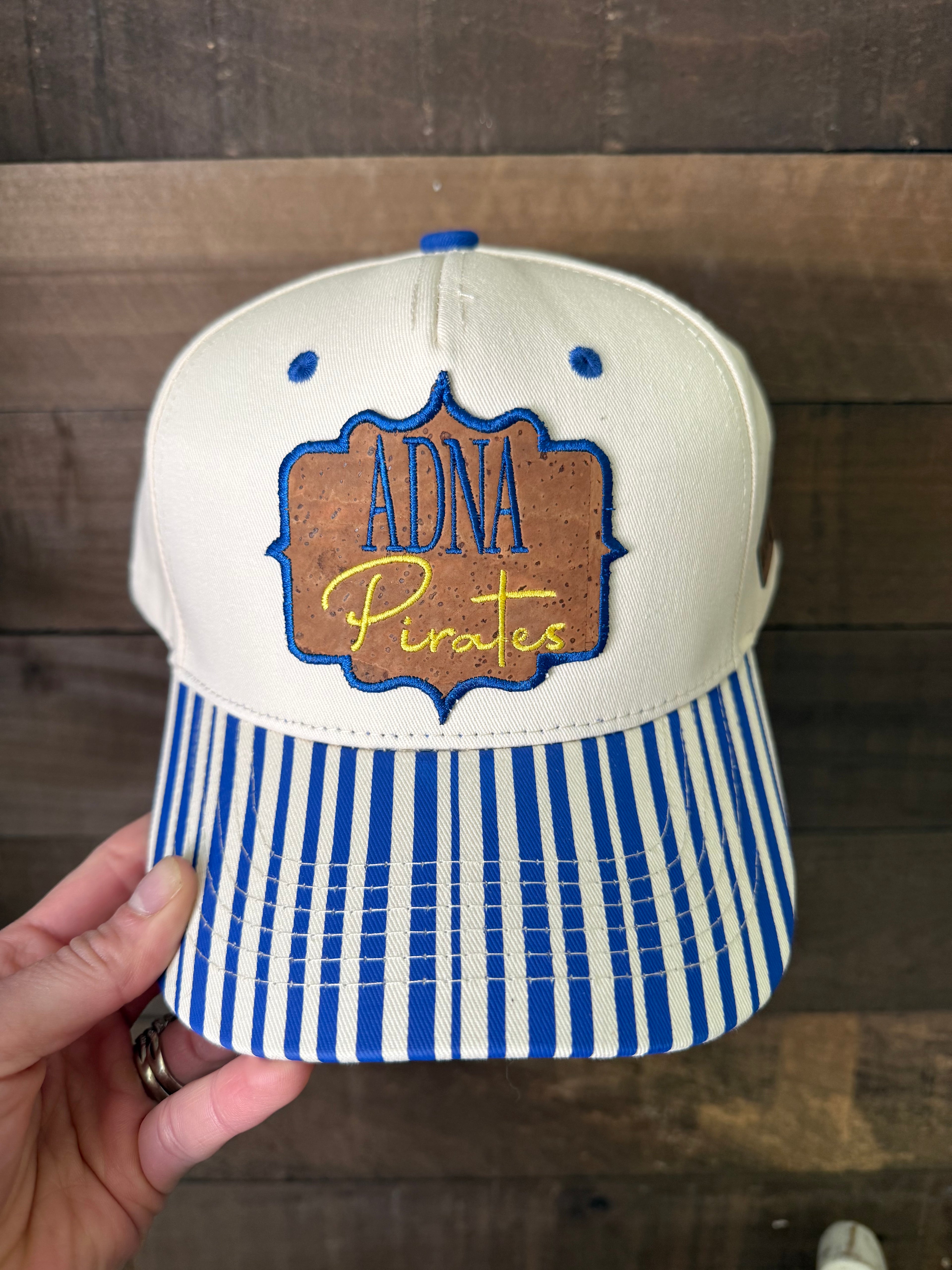 Baseball cap with 'Adna Pirates' logo held against a wooden background